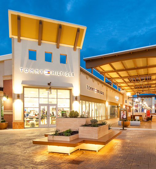 An outdoor shopping center at dusk features a Tommy Hilfiger store with illuminated signage and a seating area with planters in the foreground.