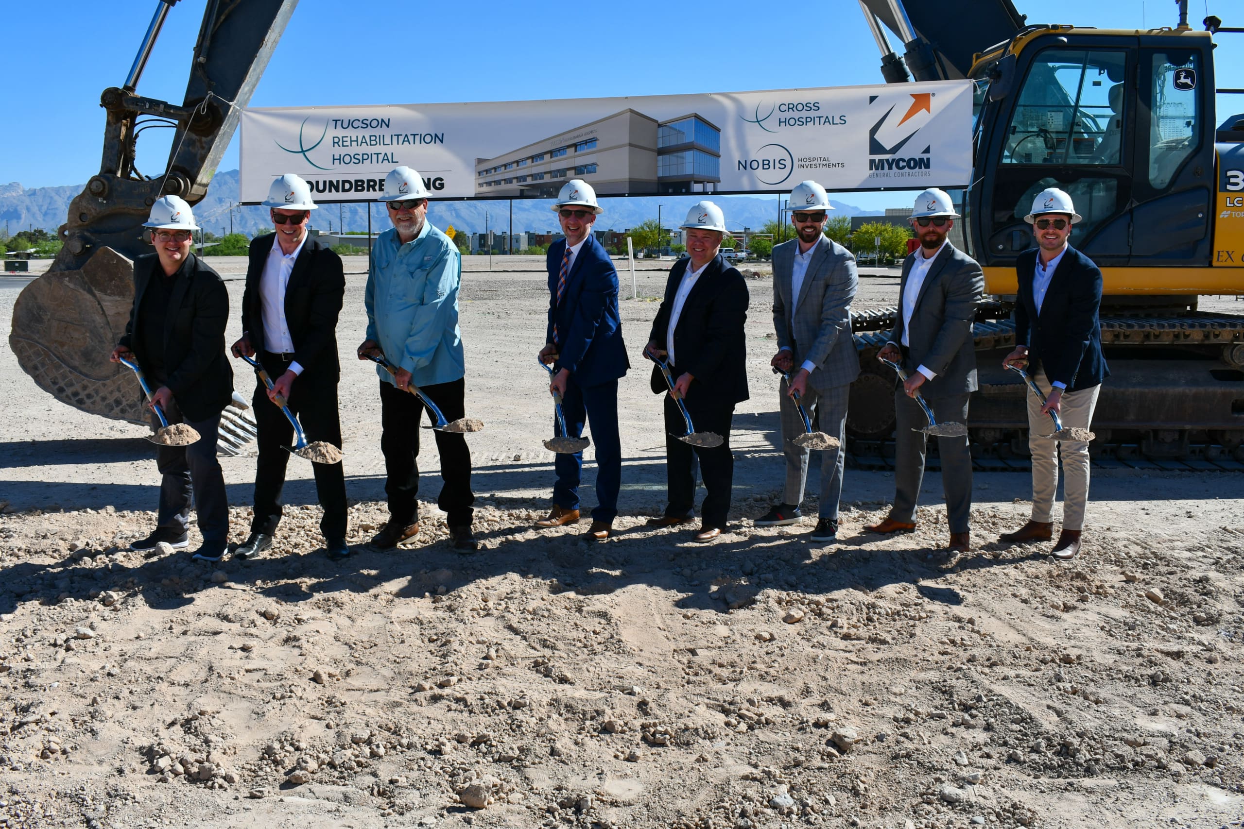 Seven people in hard hats and formal attire stand in front of an excavator, holding shovels at a groundbreaking ceremony for Tucson Rehabilitation Hospital. A banner with details of the event is behind them.