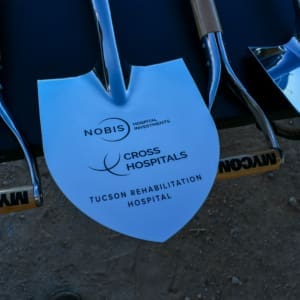 Shovels with "Nobis Hospital Investments" and "Cross Hospitals Tucson Rehabilitation Hospital" printed on the blades, lined up for a groundbreaking ceremony on a dirt surface.