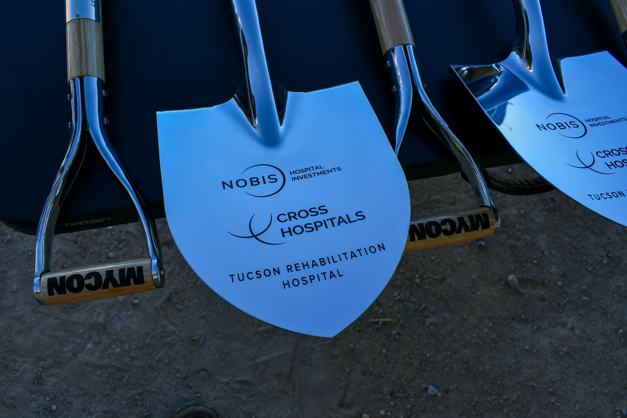 Shovels with "Nobis Hospital Investments" and "Cross Hospitals Tucson Rehabilitation Hospital" printed on the blades, lined up for a groundbreaking ceremony on a dirt surface.