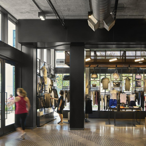 A modern retail store interior with clothing displayed on racks and shelves. Two people are shopping, and large windows allow natural light to illuminate the space.