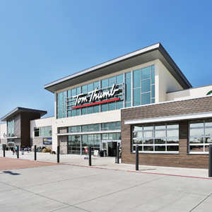 A modern grocery store exterior with large glass windows and a sign reading "Tom Thumb." Clear blue sky above and a clean pavement in front.