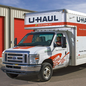 A white U-Haul moving truck is parked outside a building with red storage unit doors. The driver is visible through the window. The truck is branded with U-Haul logos and orange checkered patterns.