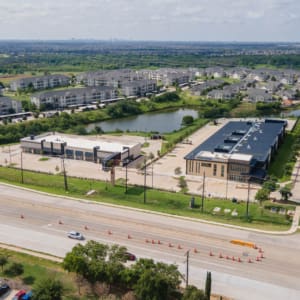Aerial view of a suburban area with commercial buildings, Parkwood Collision Center Construction, a pond, residential apartments, green spaces, and a road with traffic cones and parked cars.