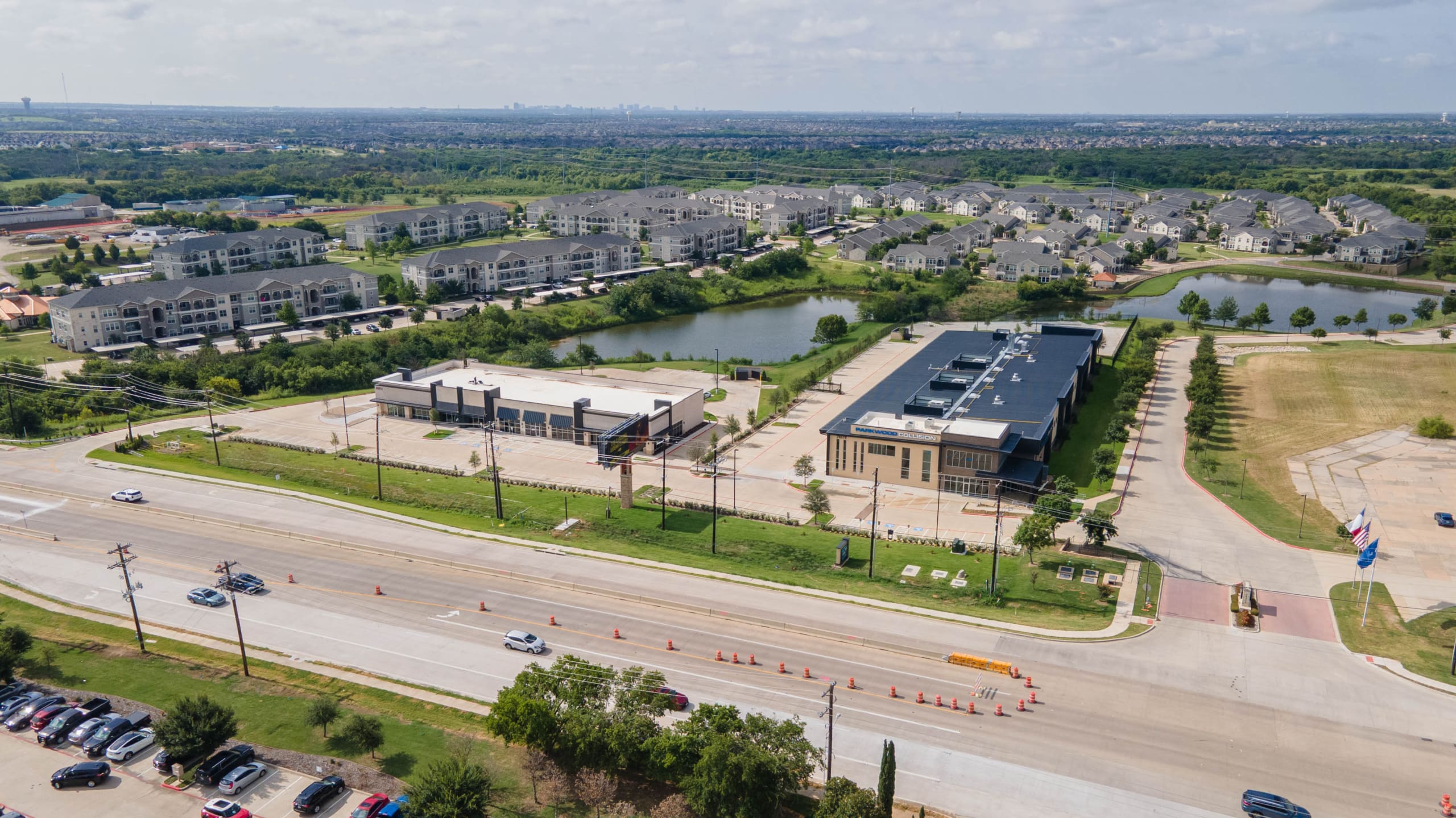 Aerial view of a suburban area with commercial buildings, Parkwood Collision Center Construction, a pond, residential apartments, green spaces, and a road with traffic cones and parked cars.