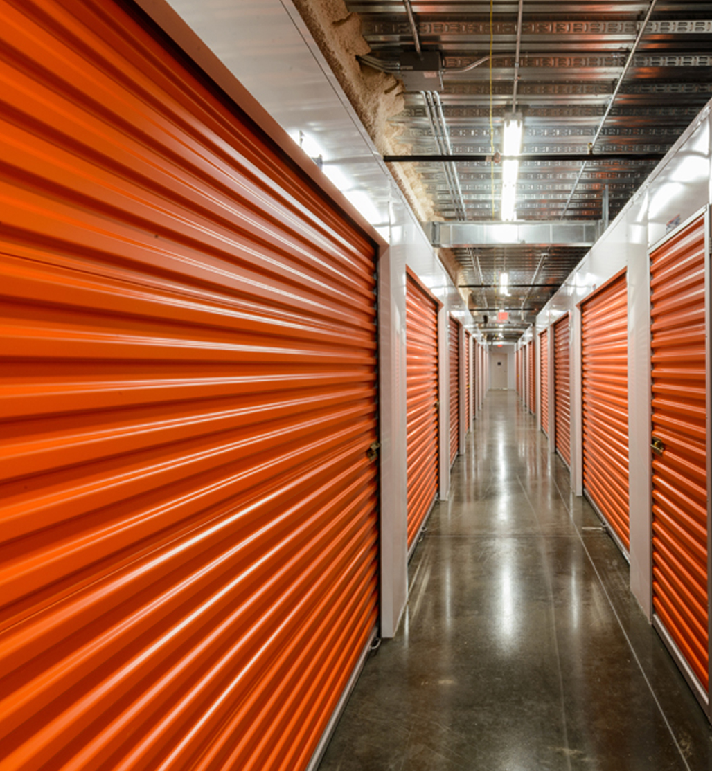 A clean, well-lit hallway lined with orange roll-up storage unit doors in an indoor self-storage facility.