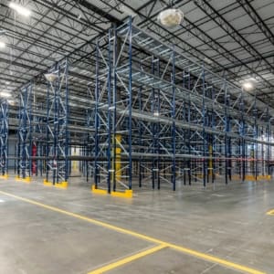 An empty industrial warehouse with metal shelving units and a high ceiling, illuminated by overhead lights. The concrete floor is marked with yellow lines.