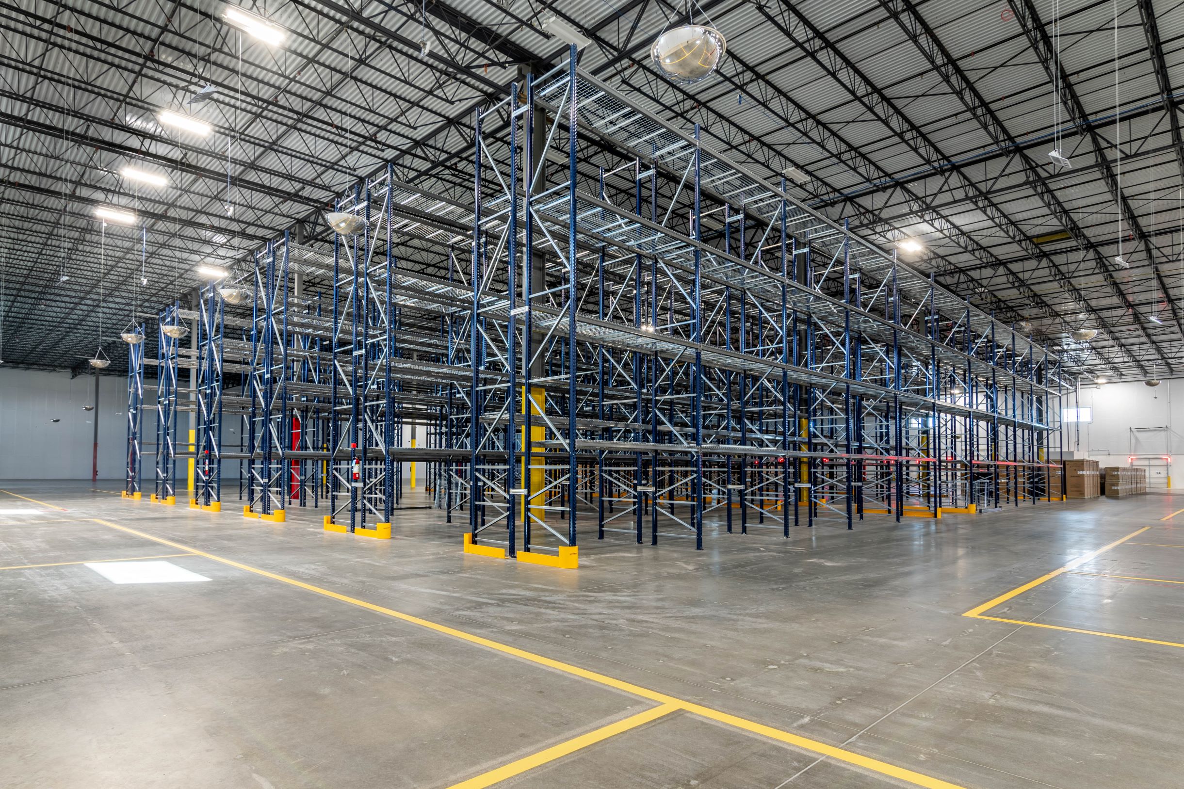 An empty industrial warehouse with metal shelving units and a high ceiling, illuminated by overhead lights. The concrete floor is marked with yellow lines.