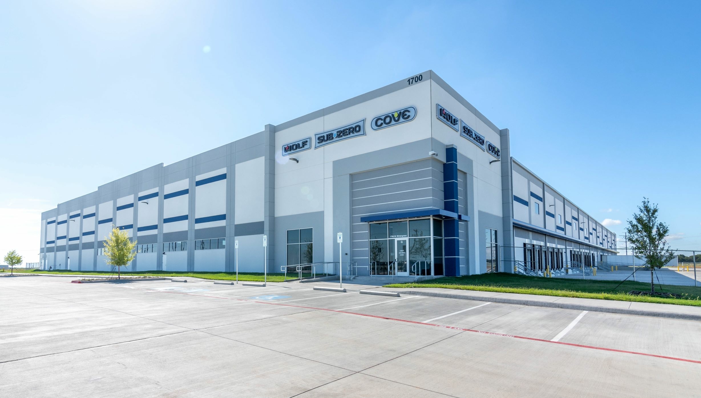 Large industrial warehouse building with company logos on the facade, situated in a paved parking lot under a clear blue sky.