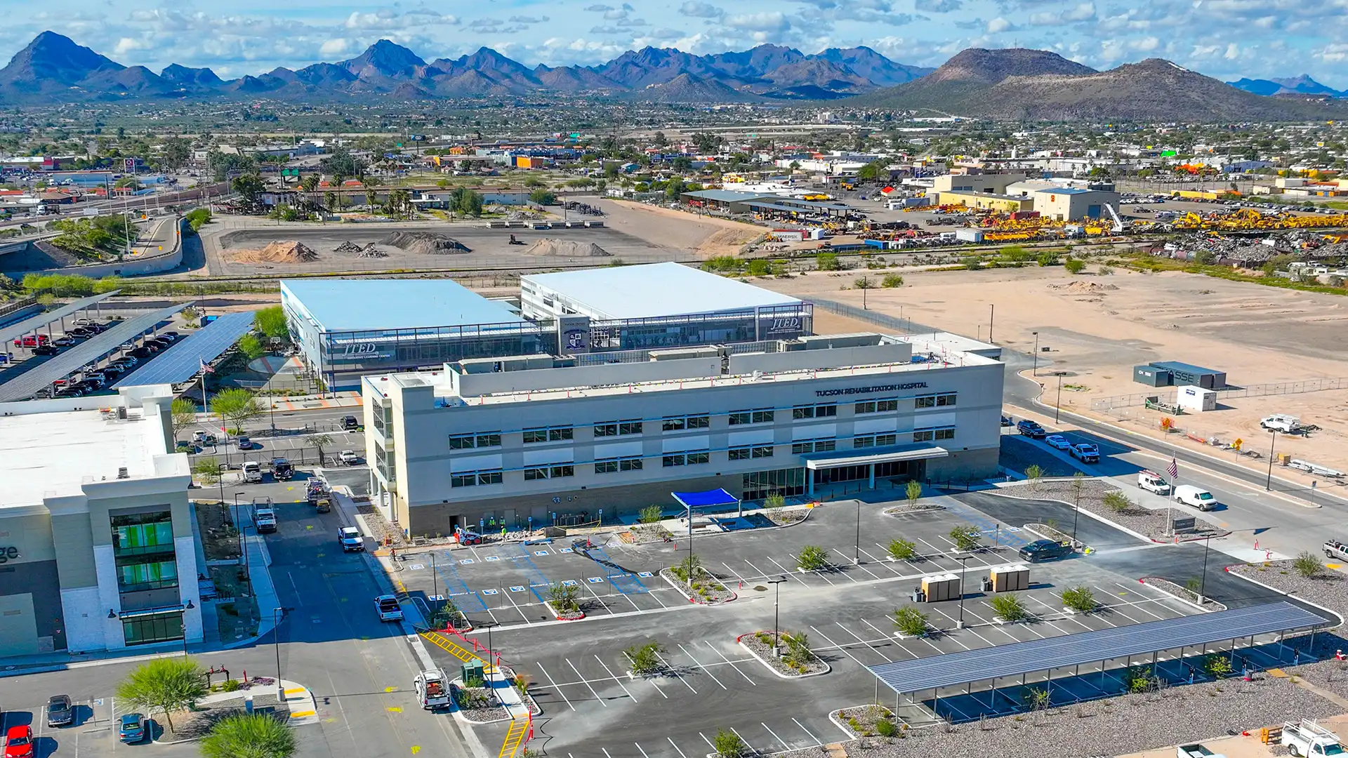 Aerial view of Tucson Rehabilitation Hospital, a modern medical building with a parking lot in a suburban area, mountains and scattered clouds in the background.