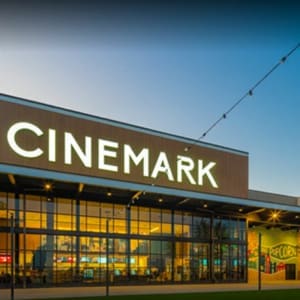Exterior view of a Cinemark movie theater with a bright sign, large glass windows displaying inside, and a popcorn-themed mural on the right side.