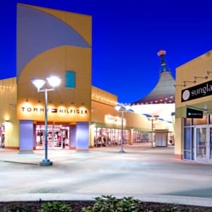 Outdoor shopping mall at dusk with stores including Tommy Hilfiger and Sunglass Hut, brightly lit with modern architectural elements and a central walkway.