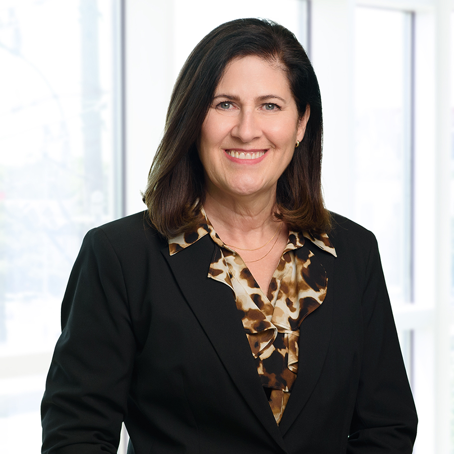 A woman with shoulder-length dark hair is smiling, wearing a black blazer over a patterned blouse. She is standing in front of a bright background with large windows.