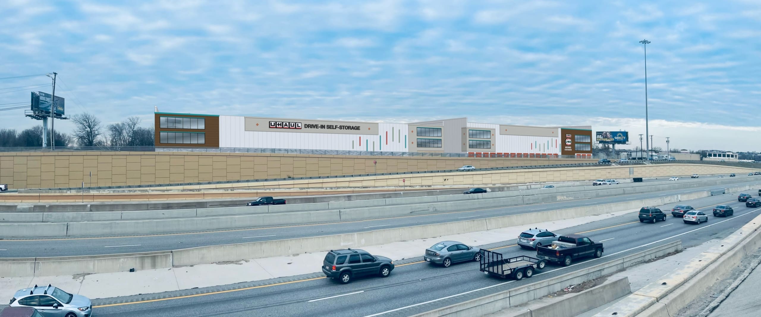 A large, modern warehouse building along a busy multi-lane highway with light to moderate traffic. The sky is overcast with streaks of clouds.