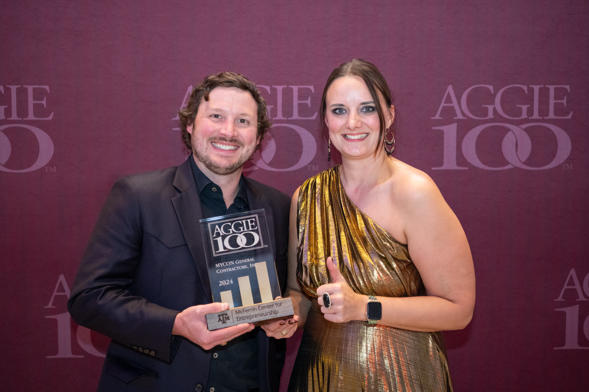 Two people pose with an Aggie 100 award. One person holds the award plaque, while the other gives a thumbs-up. They stand in front of an Aggie 100 logo backdrop.