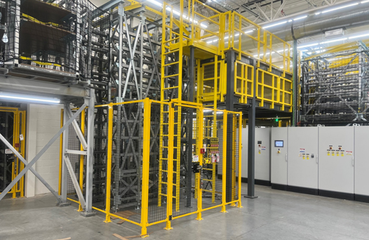 A large industrial facility with yellow safety railings, structural steel racks, and control panels. The floor is concrete, and the area is brightly lit.