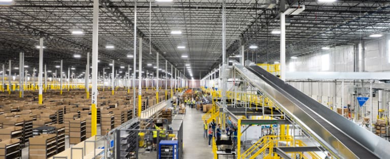 Wide view of a large warehouse with high ceilings, numerous shelves filled with boxes, a conveyor system, and workers moving between aisles.