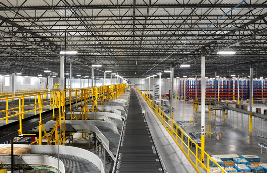 Interior of a large distribution warehouse with conveyor belts, yellow railings, and tall racks filled with goods, under a high ceiling with a grid of metal beams and bright lighting.