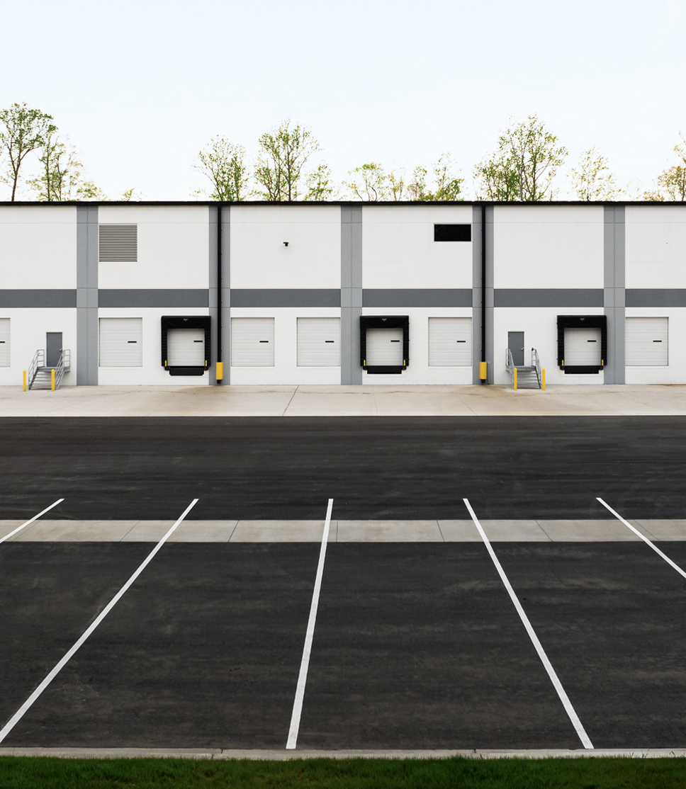 Warehouse building with four loading docks and a large, empty parking area in front. Sparse trees are visible above the building.