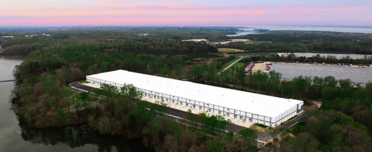 A large, rectangular white warehouse surrounded by trees and water, set against a backdrop of a sunset sky and distant hills.