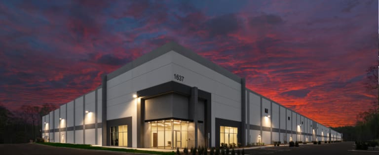 Large warehouse building with exterior lights on, marked "1637," set against a vivid sunset sky.