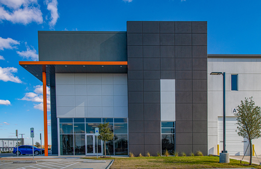 Modern building exterior with a geometric design, featuring gray and white panels, an orange accent, large glass windows, and a small tree in front. A blue car is parked to the left.