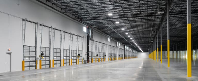 Interior of an empty, expansive warehouse with high ceilings, yellow safety bollards, and closed garage doors lining one wall.