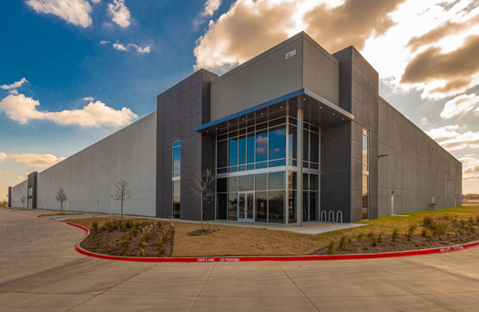 A large, modern industrial building with a gray exterior, glass entrance, and surrounding pavement under a partly cloudy sky.