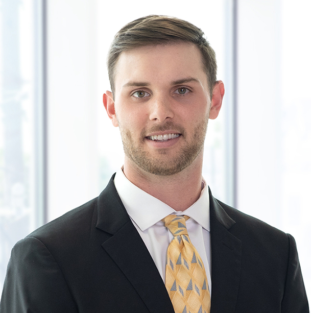 A man in a suit with a patterned yellow tie stands in front of a windowed background.