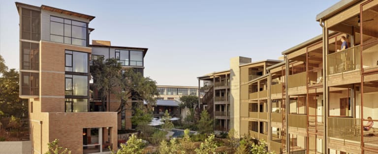 A modern apartment complex with multiple buildings and greenery in the courtyard under a clear sky.