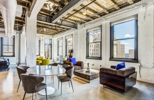 A modern living area with large windows, exposed beams, and a mix of seating, including chairs around a round table and cushioned sofas. Bright natural light fills the space.