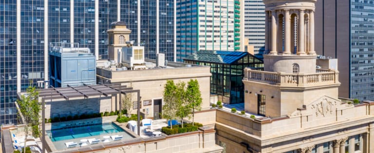 Rooftop with a swimming pool, lounge chairs, and greenery, surrounded by tall city buildings.