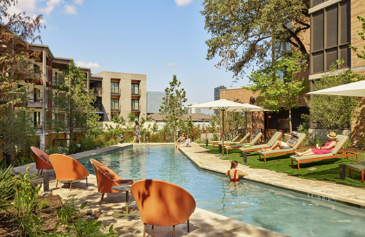 Outdoor swimming pool with people relaxing on loungers under umbrellas. Surrounded by trees and modern buildings in the background.