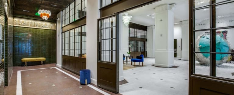 Spacious modern lobby with green-tiled walls, glass partitions, and a chandelier. A blue chair and small table are in the seating area. Large decorative glass display on the right wall.