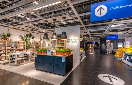 Furniture showroom with modern designs, hanging lights, and potted plants. Arrow signs on the ceiling guide the flow of visitors.