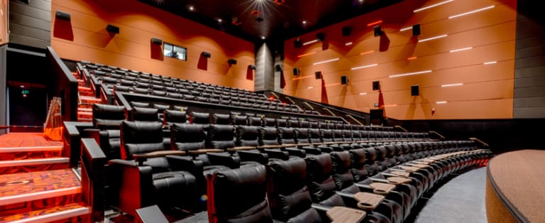 An empty movie theater with rows of black reclining seats and armrest tables, orange walls with rectangular lights, and a patterned carpeted floor.