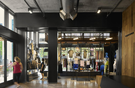 A modern clothing store interior with racks of clothes and mannequins. A person walks through the entryway on the left. The decor includes black and wooden elements.