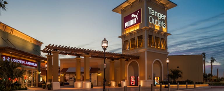 Tanger Outlets entrance at dusk, featuring Puma and Under Armour stores with illuminated signage and architectural details.