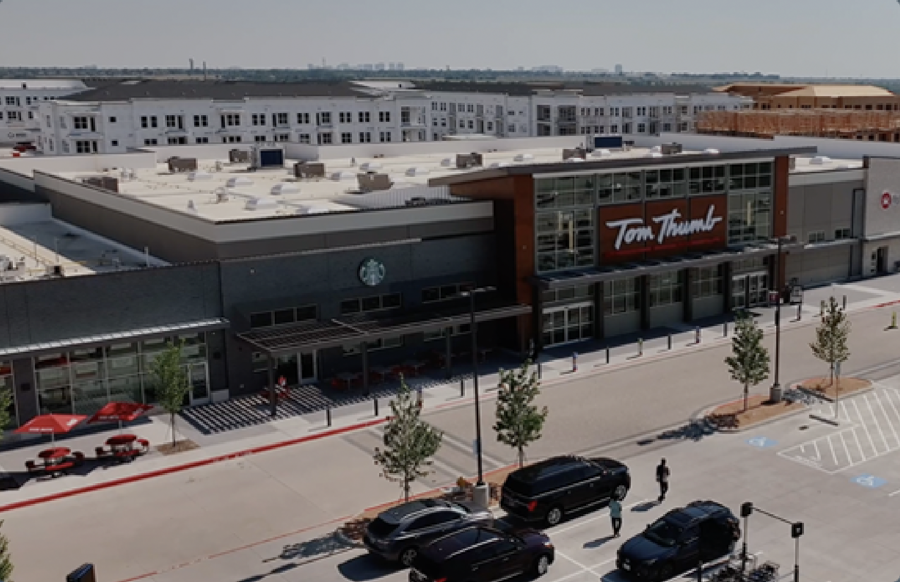 Aerial view of a grocery store with "Tom Thumb" signage, located in a commercial area with adjacent parking and nearby apartment buildings.