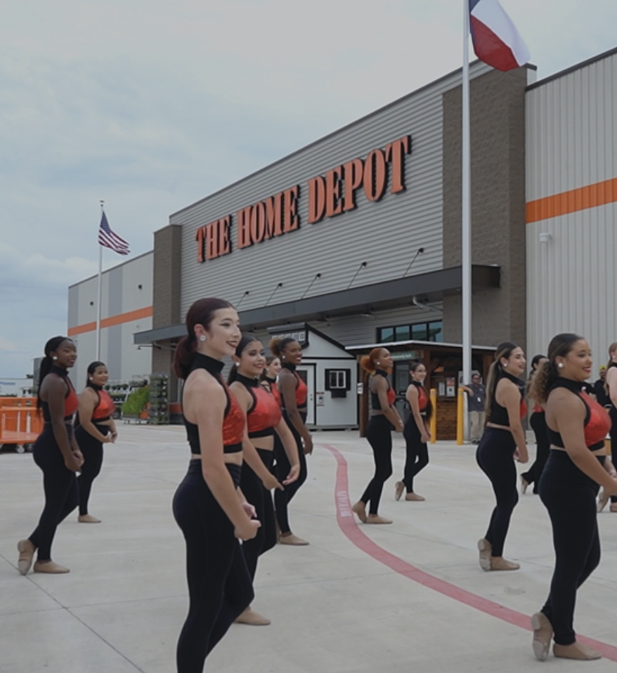 A group of dancers in black outfits and red tops perform outside an entrance of The Home Depot, with American and Texas flags visible.