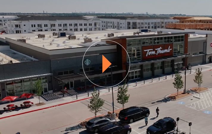 Aerial view of a Tom Thumb grocery store with a Starbucks logo on the side. Parking lot in the foreground, and a building complex in the background under a clear sky.