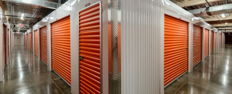 A hallway with several orange roll-up doors of storage units on a polished concrete floor. Ceiling features exposed ducts and lights.