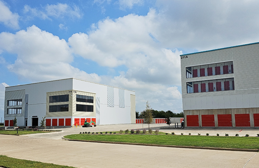 Two industrial buildings with white and red exteriors under a partly cloudy sky. A paved road and grassy area are in the foreground.