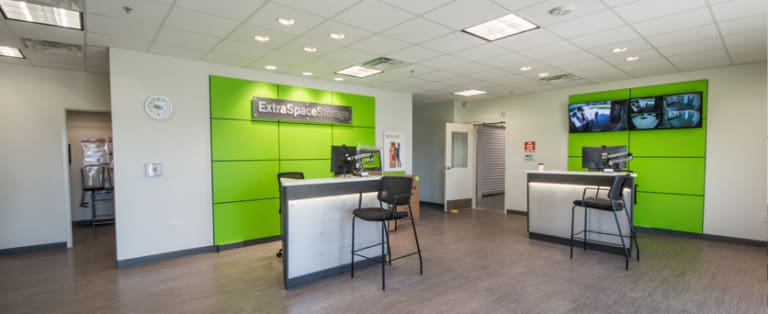 A modern storage facility office with green and white decor, featuring a reception desk, chairs, monitors, and security camera screens on the wall.