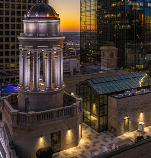 Rooftop view of a cityscape at dusk with a domed tower, illuminated building facades, and a glass-roofed structure, under a colorful sunset sky.