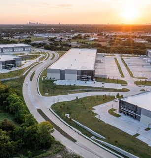 Aerial view of a business park with several large warehouse buildings, surrounded by roads and green spaces, under a setting sun.