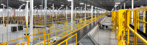 Interior view of a large warehouse with extensive conveyor systems, bright overhead lighting, and numerous storage racks filled with boxes and packages.