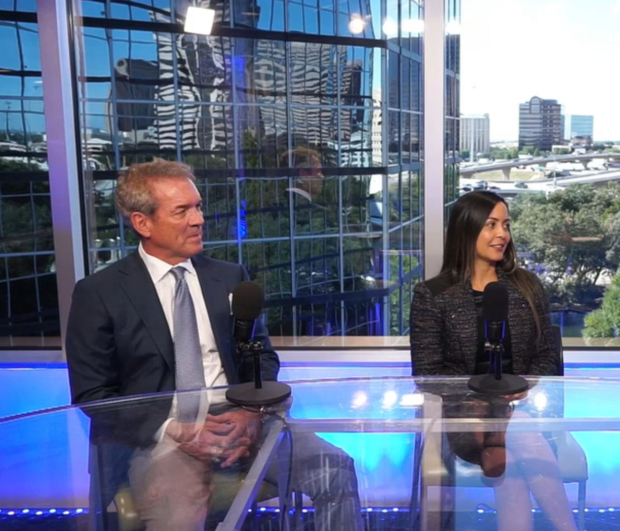 Three people in business attire sit at a glass table with microphones, engaged in conversation in a studio with cityscape windows behind them.