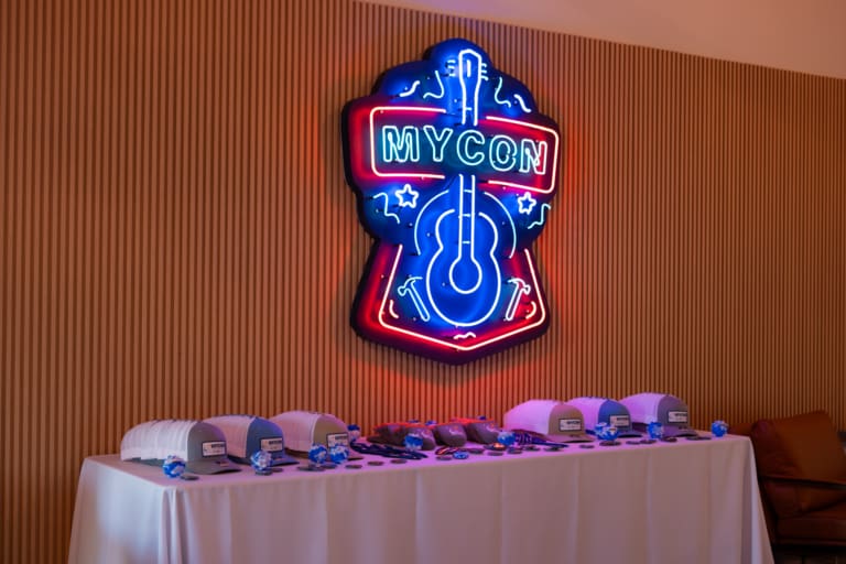 Table with hats and lanyards below a neon sign reading "MYCON," featuring a guitar and music notes.