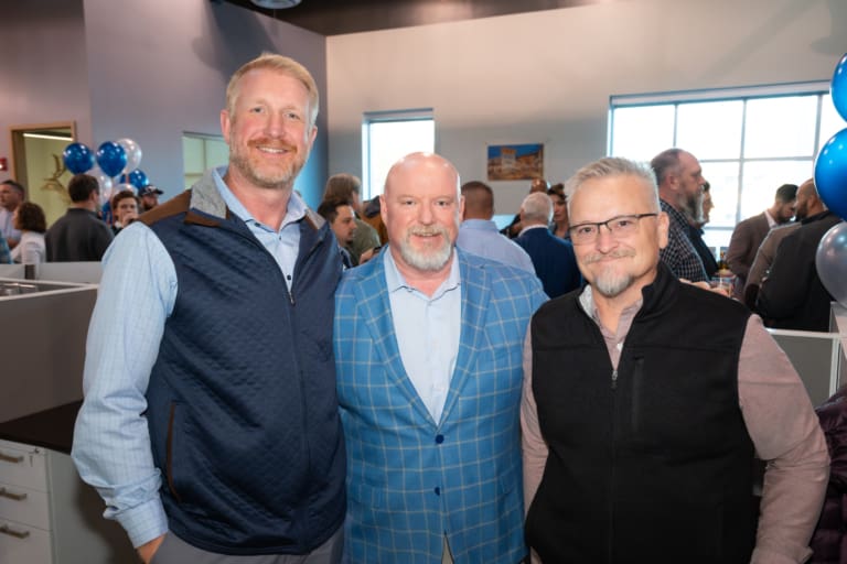 Three men standing together in an office surrounded by people and blue balloons.